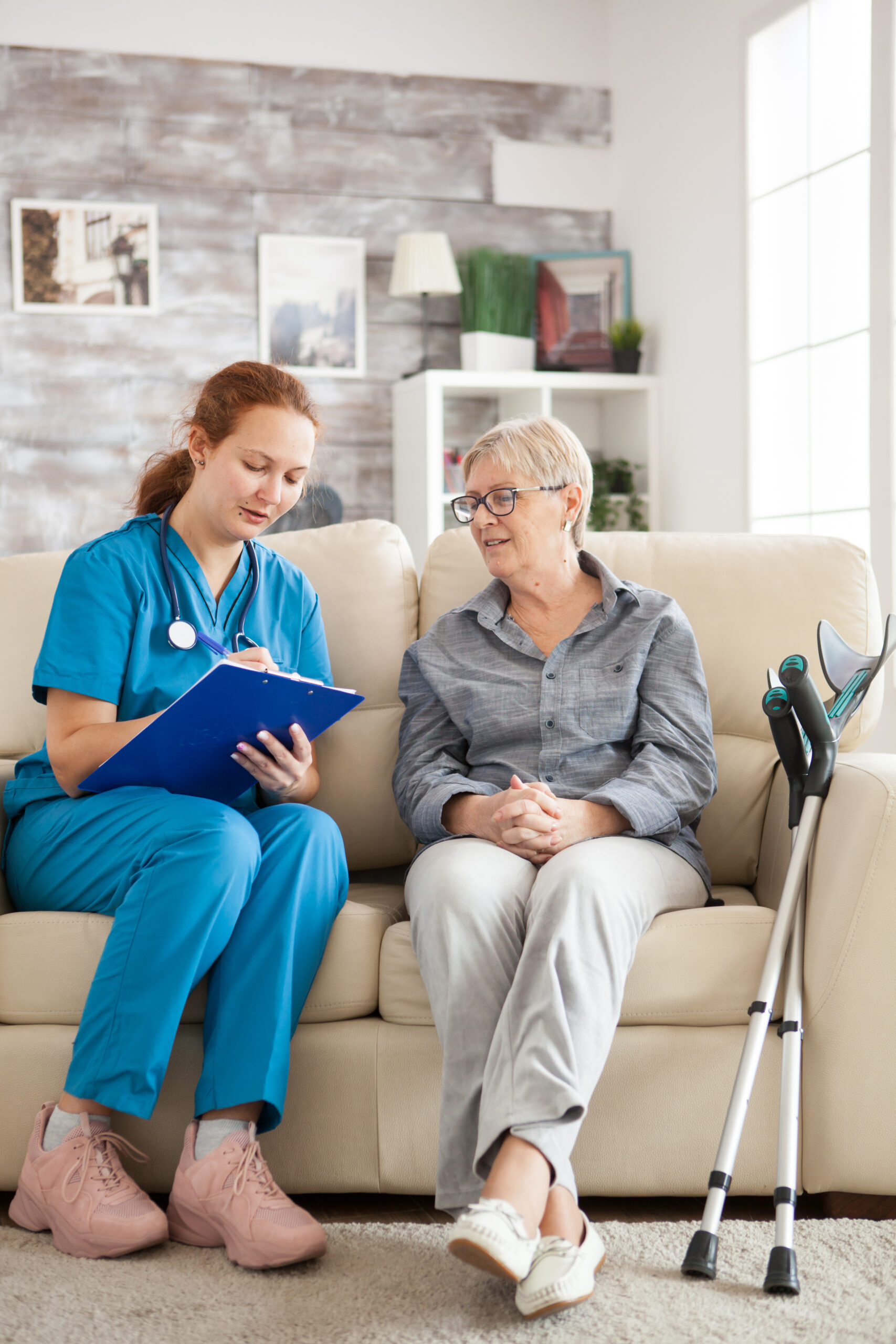 Female doctor sitting on a couch with a senior woman in a nursing home, writing a prescription on a clipboard.